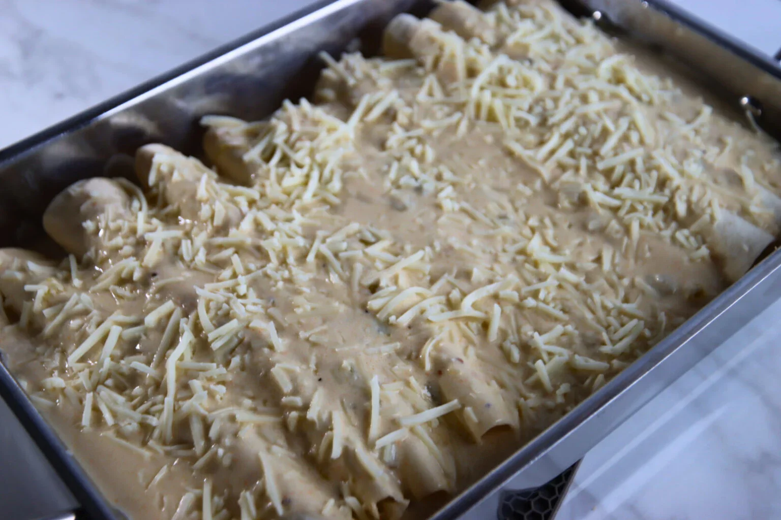 Rolled chicken enchiladas covered with creamy white sauce and shredded Monterey Jack cheese in a baking pan.