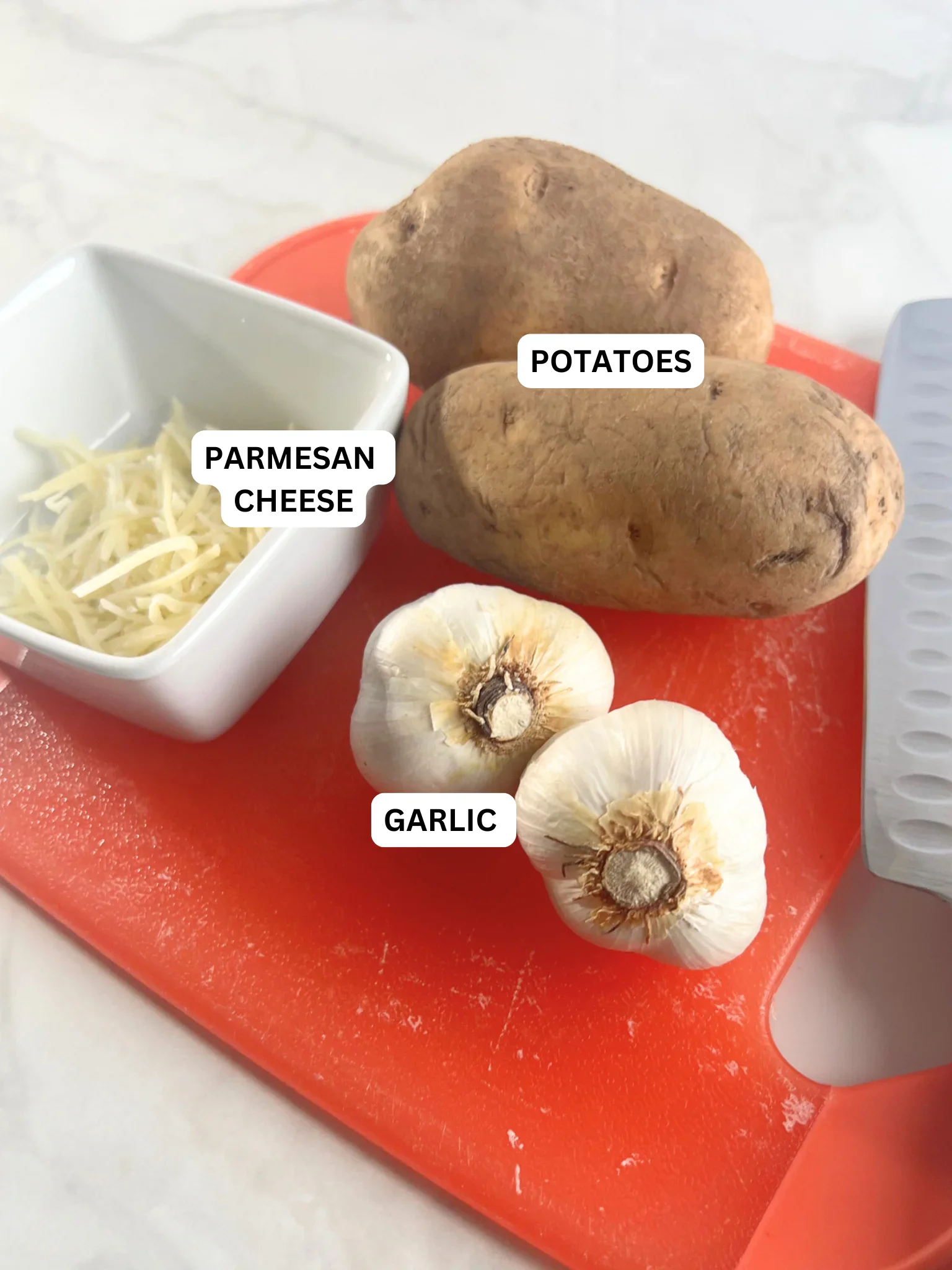 Russet potatoes, garlic bulbs, and shredded Parmesan cheese on a cutting board.